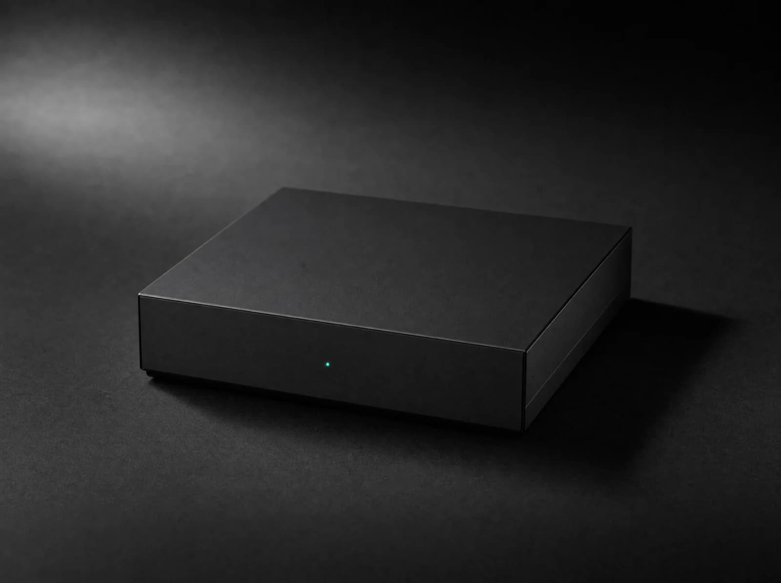 A compact unbranded matte-black appliance on a dark surface, lit from one side, with a single teal status indicator on its front face.